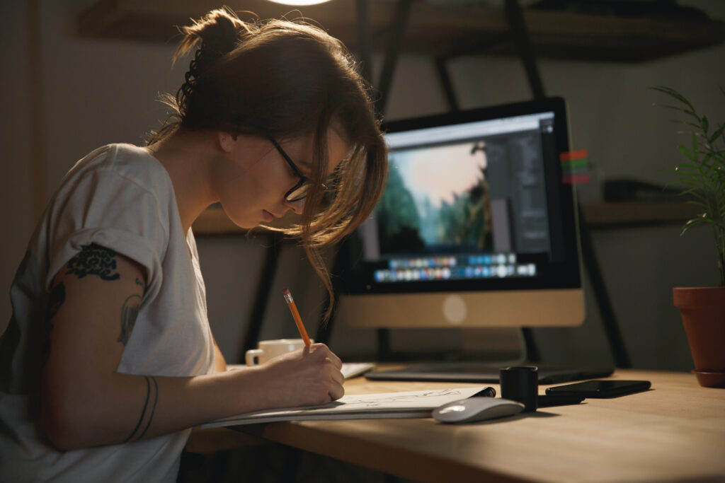 Photo of young woman designer sitting indoors at night drawing sketches in album while using computer.